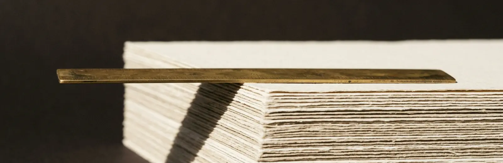A brass rule resting across a stack of bone-coloured paper in raking light